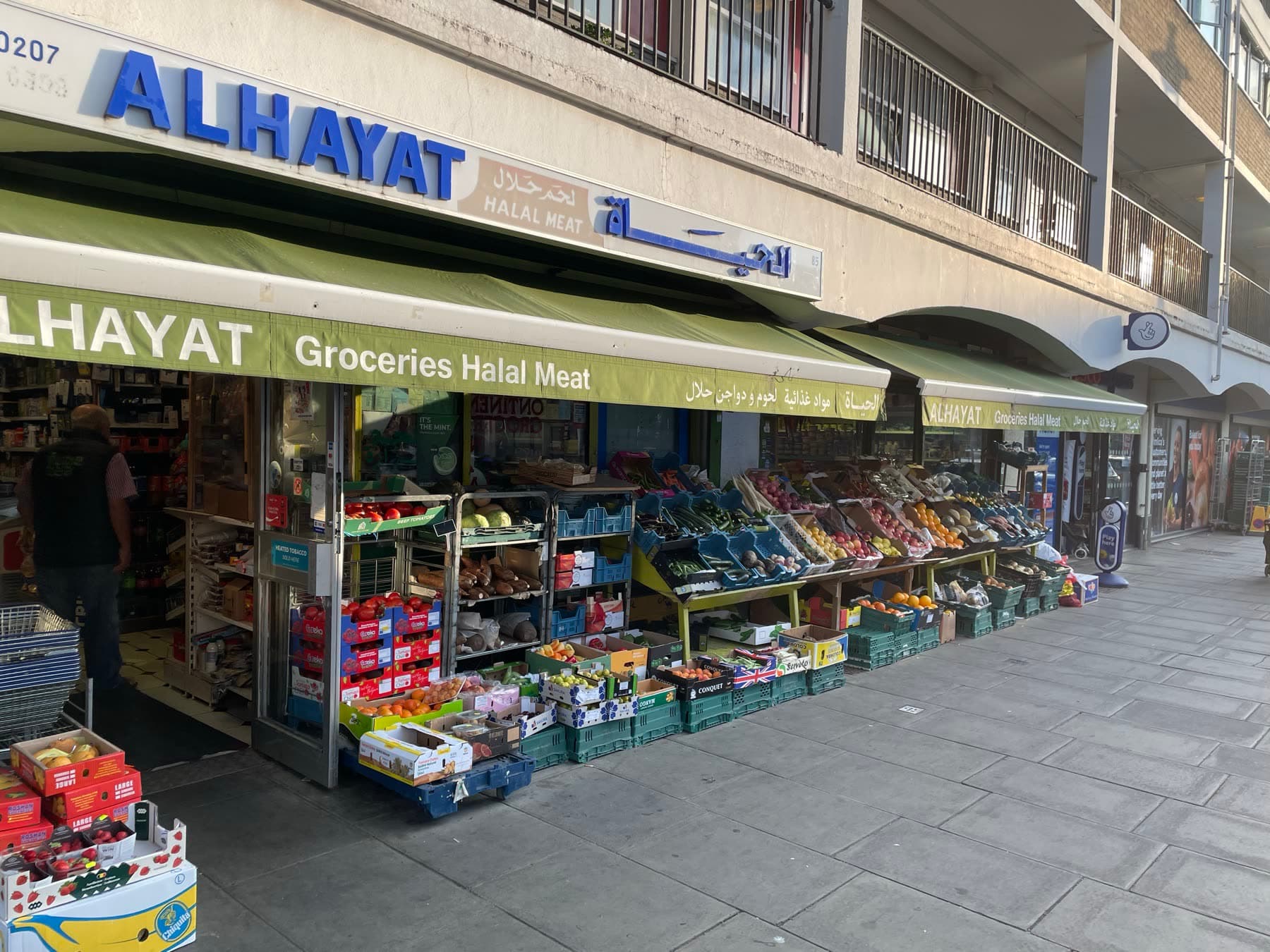 The full run of the Alhayat shopfront along the pavement, produce crates set out beneath three green awnings